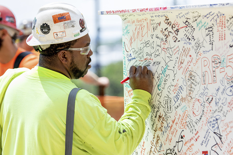 A member of the PennFIRST construction team wearing a yellow vest and hard hat adds his signature to the final beam before it is placed atop the Pavilion structure.